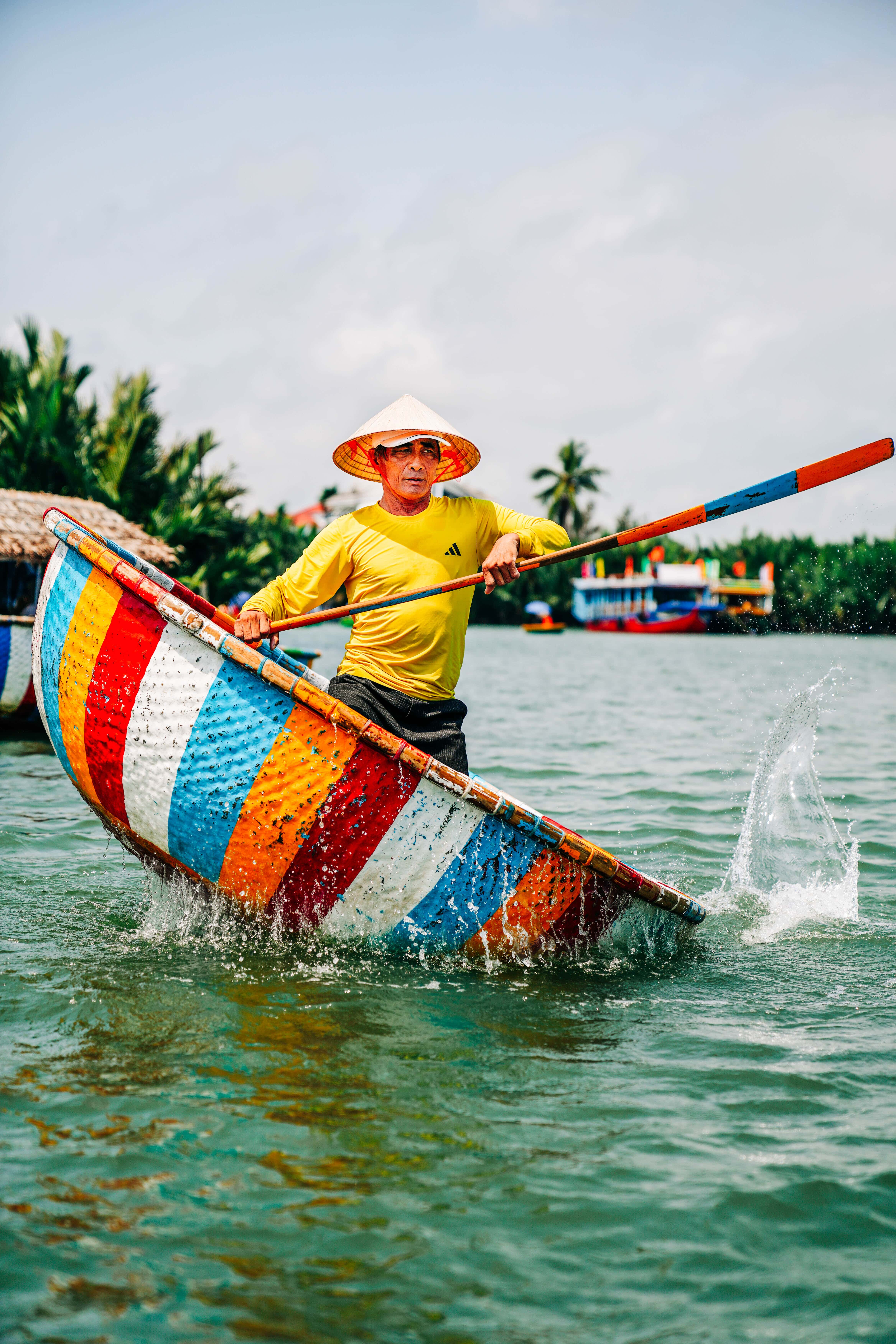 A man in a colorful boat. The boat is round resembling a coconut shape. The man is aggressively paddling and the water is splashing around him.