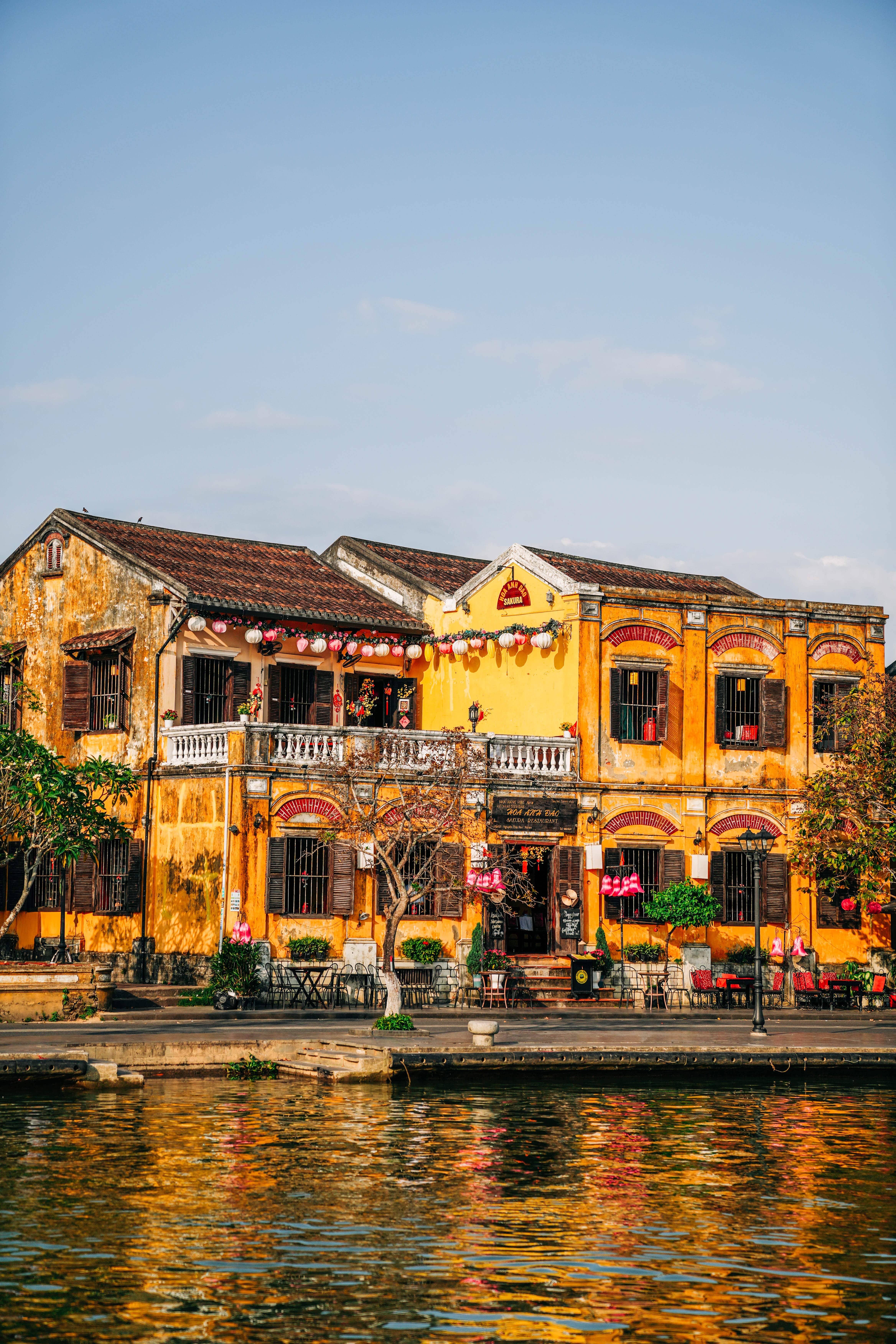 A brightly colored yellow building with arches and terraces. Lanterns hung on trees. The colors are reflecting on the water in front of the structure.