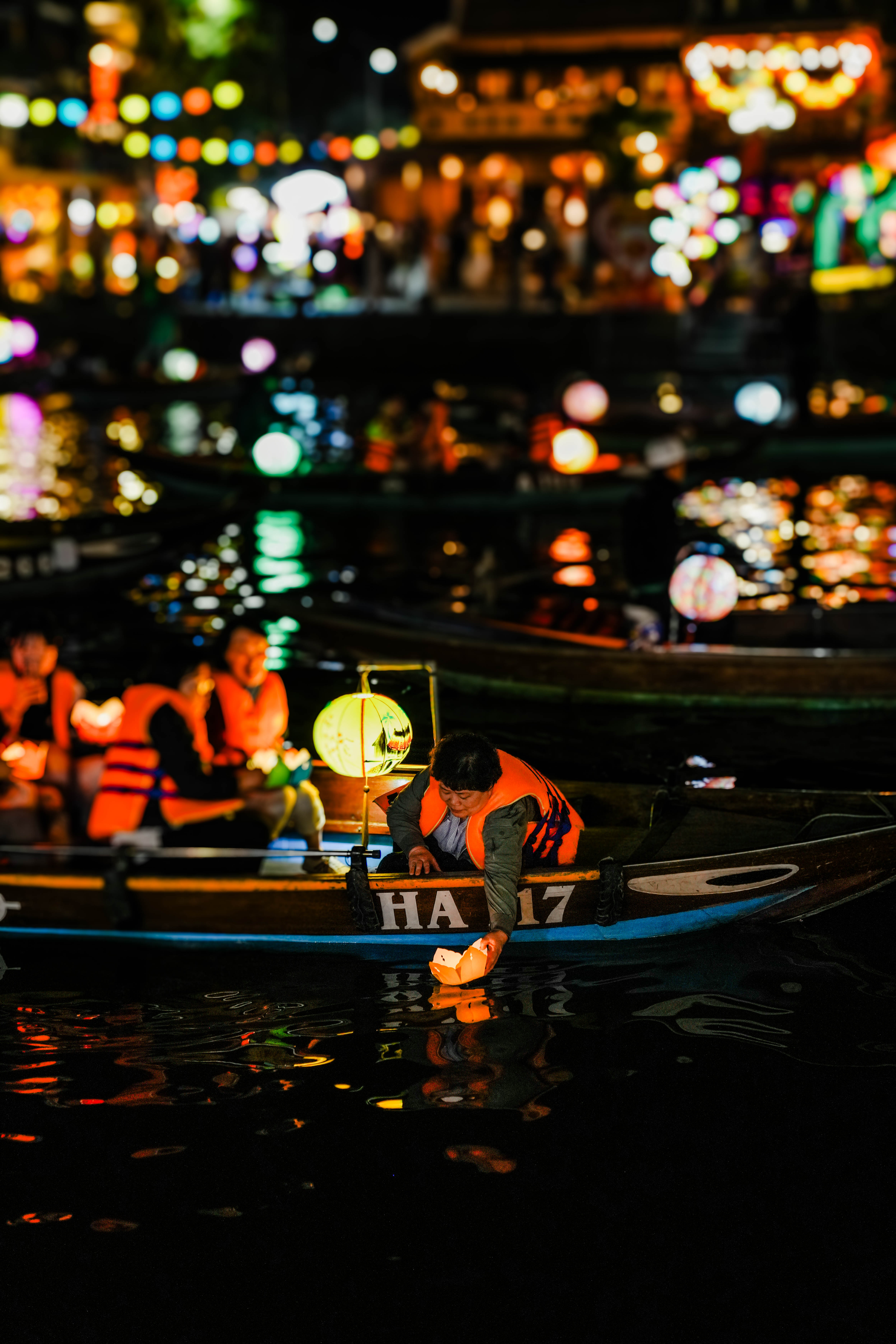 A woman in a boat with others. She is releasing a lantern into the water. Lights glow in the water's reflection.