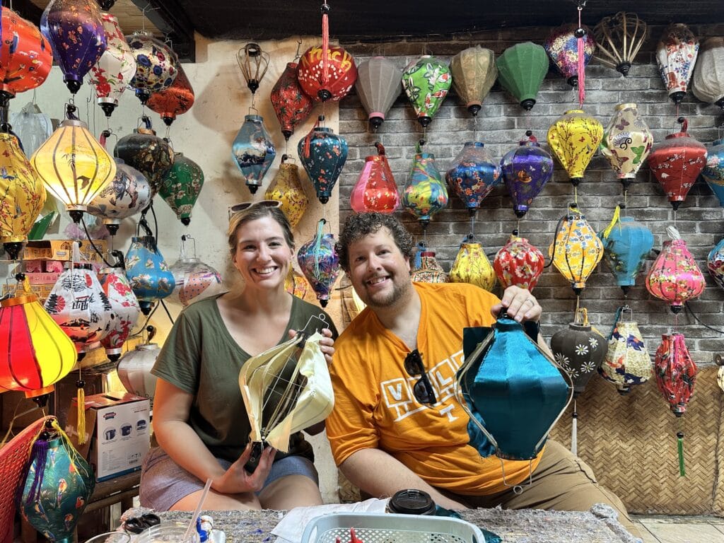 Two people posing for a photo in front of a wall of hanging lanterns. The couple are holding a lantern each.